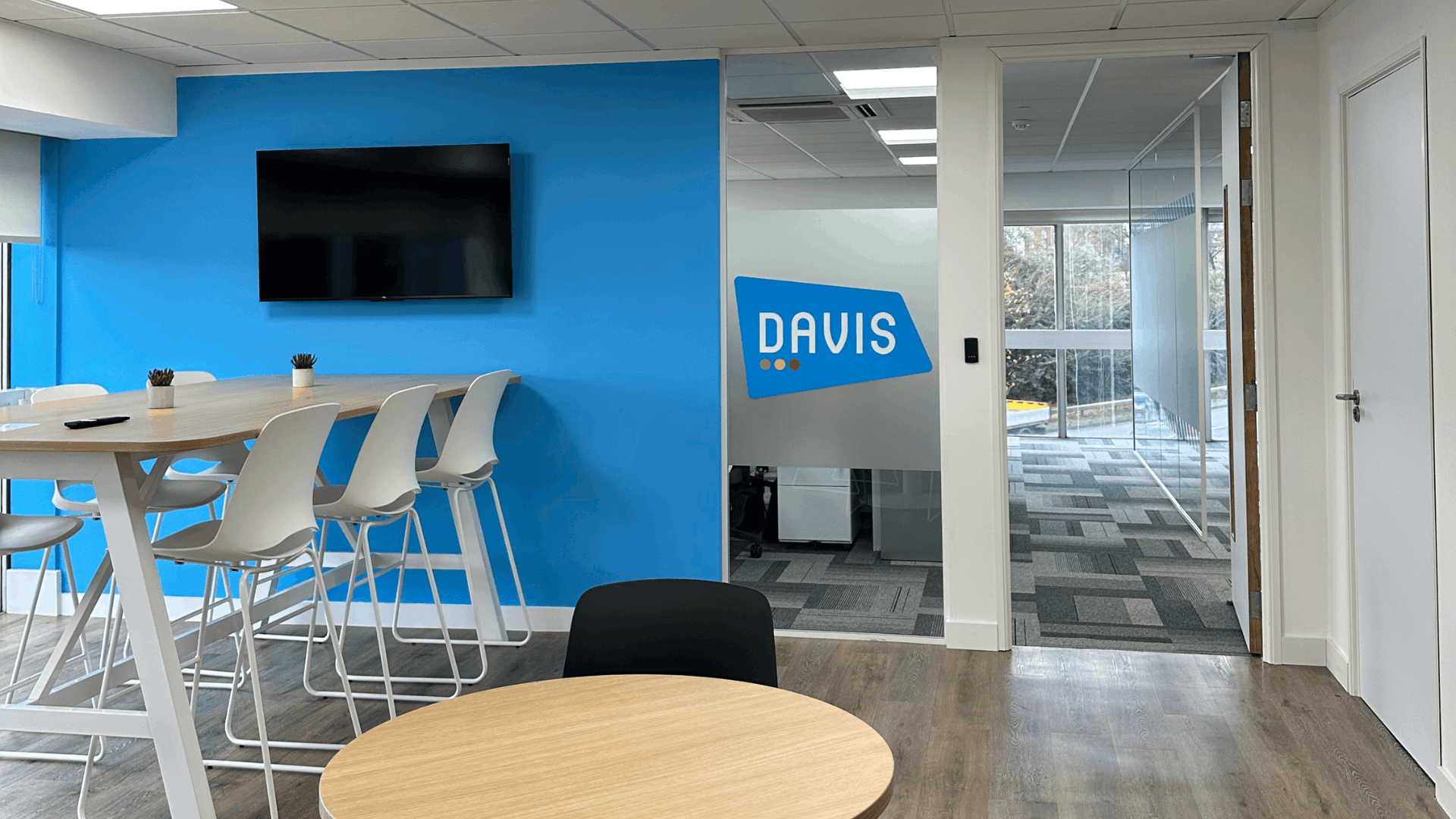 Completed License Check office fit out breakout area with blue feature wall, wall-mounted TV, high bench seating, round tables, wood-effect flooring and glazed office partitions